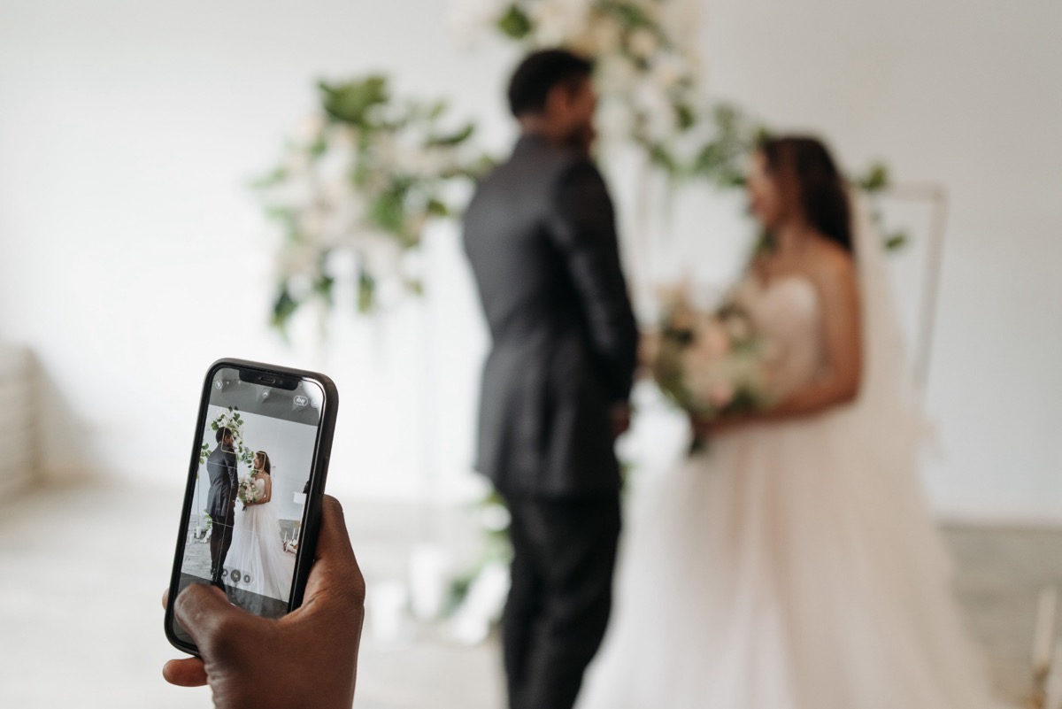 Alguien toma foto con su celular a los novios frente a un arco de flores